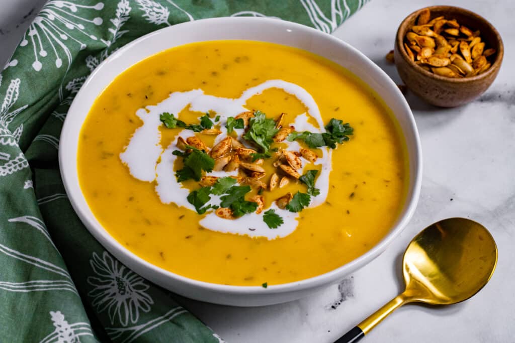 White bowl of butternut squash soup with a green napkin and some roasted pumpkin seeds.
