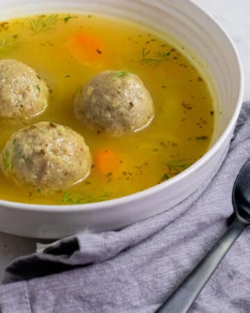 3 matzo balls and broth in a white bowl with a black spoon and a gray napkin.