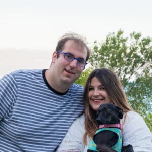 Paul and Liz holding a small black dog standing in front of Lake Michigan