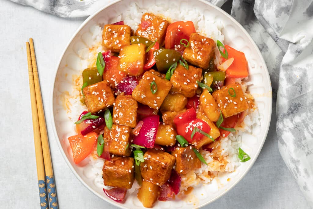 Colorful sweet and sour tofu on top of white rice in a large bowl on a white counter with wooden chopsticks