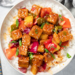 Colorful sweet and sour tofu on top of white rice in a large bowl on a white counter with wooden chopsticks