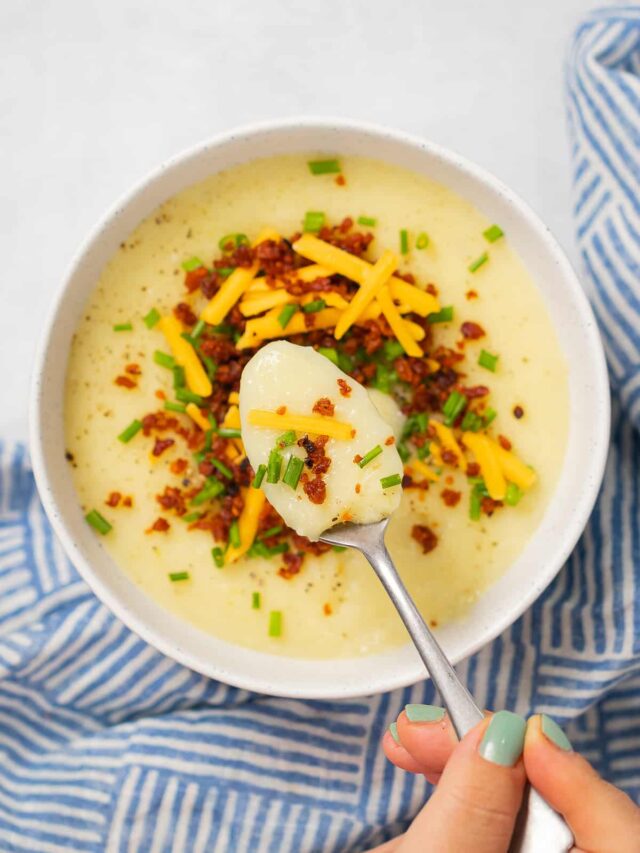 Spoon lifting a bite of vegan potato soup over a white bowl and blue striped napkin.