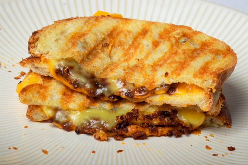 A vegan bacon grilled cheese sandwich cut in half and stacked on a white plate with melty vegan cheese.